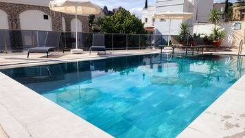 A rectangular swimming pool with clear blue water, surrounded by a tiled deck. There are lounge chairs and umbrellas, and a stone wall with arched windows in the background.