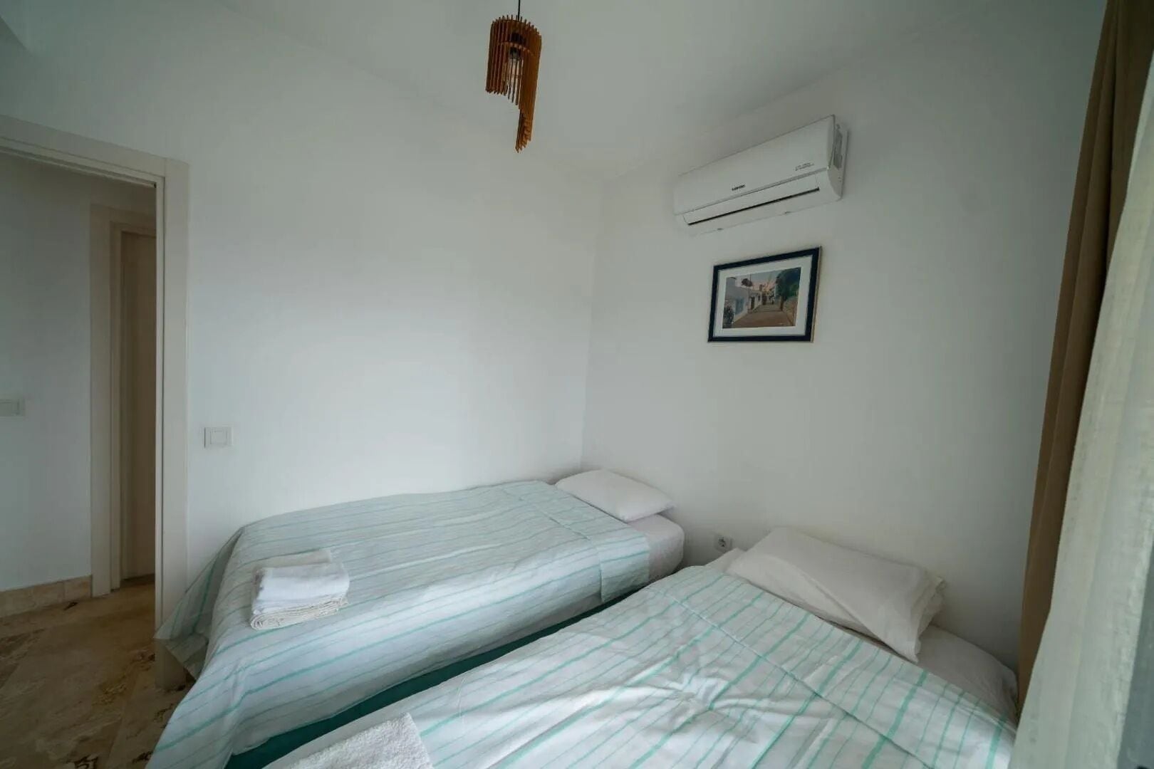 A small, clean room with two single beds, a ceiling fan, an air conditioning unit, and a framed picture on the wall.