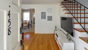 A modern living room with a staircase, wooden flooring, a flat-screen TV, and a dining area in the background. - JadeHome -- Townhome style (Alameda)