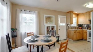 A dining area with a round table set for four, surrounded by chairs. There is a kitchen area with wooden cabinets and a countertop visible in the background. - JadeHome -- Townhome style (Alameda)