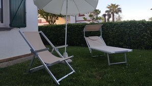 Deux fauteuils de jardin, placés sur une pelouse verdoyante, sont agrémentés d’un parasol blanc. On aperçoit un arbuste et un bâtiment en arrière-plan.