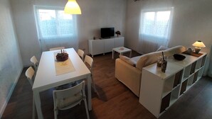 Un espace de vie compact comprenant une table à manger, deux chaises, un canapé, un petit téléviseur posé sur un meuble blanc et une lampe sur une table d’appoint.