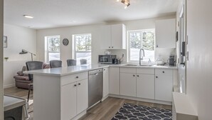 Una cocina moderna con armarios blancos, microondas integrado y lavavajillas. Hay una ventana que proporciona luz natural y una alfombra estampada en el suelo.