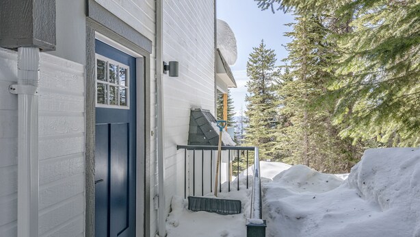Exterior nevado de una casa con puerta azul, barandilla de metal y una pala de nieve apoyada en la pared.