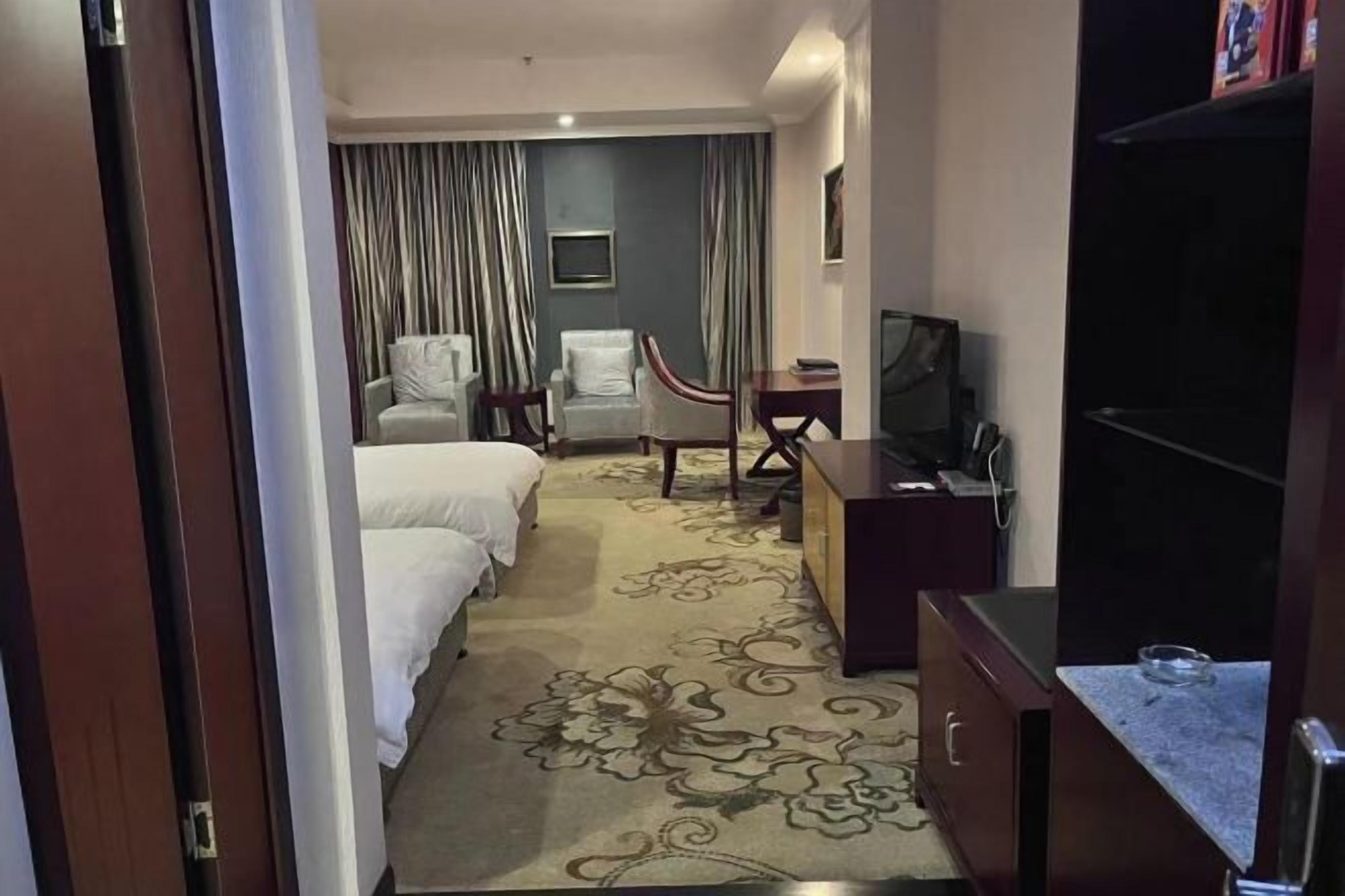 Une chambre d’hôtel avec deux lits, un bureau, une chaise, une télévision et une moquette à motifs floraux.