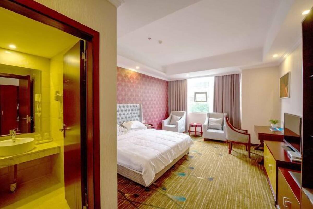 A hotel room with a bed, a desk, a chair, a TV, and a bathroom with a mirror and sink.