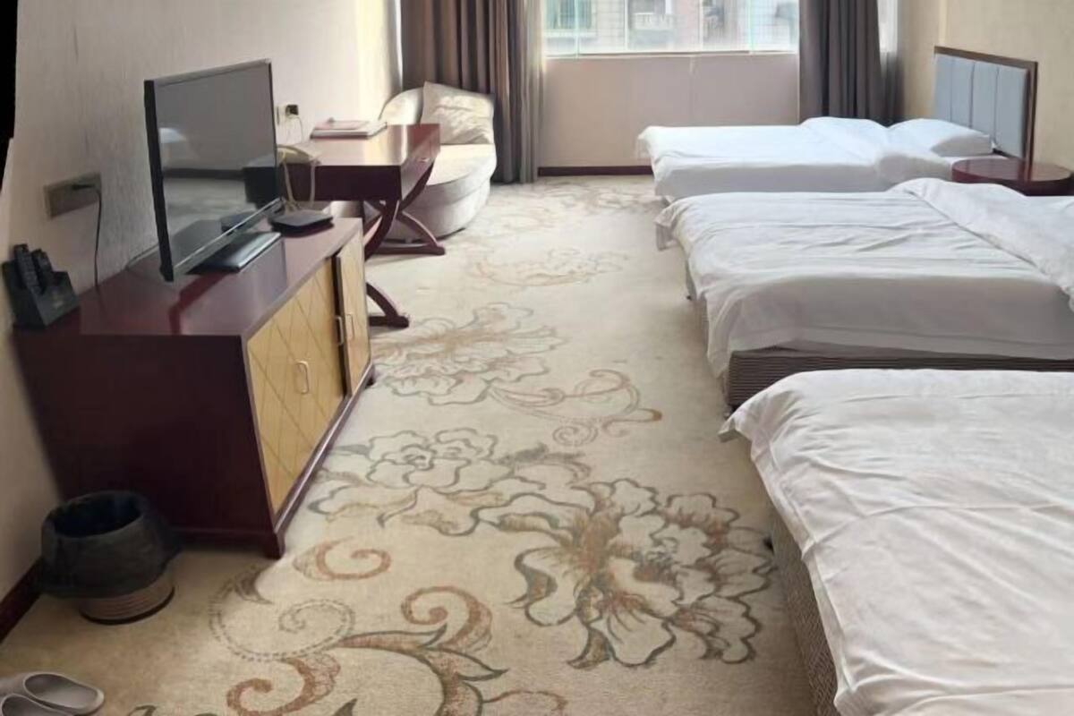 A hotel room with a flat-screen TV, a desk, two beds, a patterned carpet, and a window with curtains.