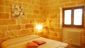 Une pièce avec un mur en pierre, un lit avec un cadre en métal blanc, une fenêtre avec des volets en bois et un radiateur.