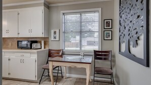 Une cuisine avec des meubles blancs, un four à micro-ondes, un grille-pain et une table à manger avec deux chaises. Il y a une fenêtre avec des stores et un panneau décoratif sur le mur, à droite.