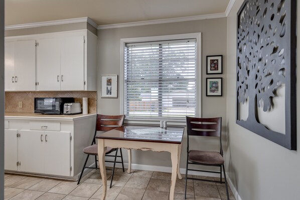 Une cuisine avec des meubles blancs, un four à micro-ondes, un grille-pain et une table à manger avec deux chaises. Il y a une fenêtre avec des stores et un panneau décoratif sur le mur, à droite.