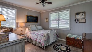 Une chambre à coucher comprenant un lit, des tables de chevet, une commode, un ventilateur de plafond et une fenêtre avec des stores.