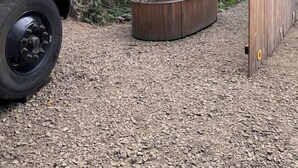 A yellow vehicle parked on gravel, a hot tub, and a wooden fence in a wooded area.