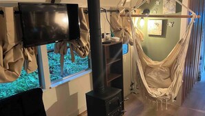 A room with a television, a black wood-burning stove, a hammock, and a window with a view of trees.