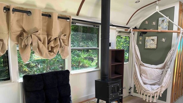 A cozy room with a black sofa, a wood-burning stove, a hammock, and large windows with curtains.