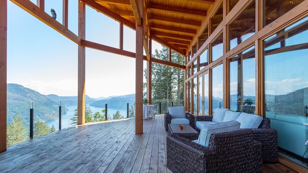 A wooden deck with wicker furniture overlooking a lake and mountains. - The Summit Room suite  – Breathtaking Fjord View at the Summit House. (Malahat)