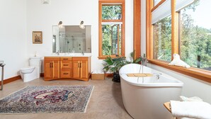 A modern bathroom with a freestanding bathtub, a large mirror, and a window with a view of trees. - The Summit Room suite  – Breathtaking Fjord View at the Summit House. (Malahat)