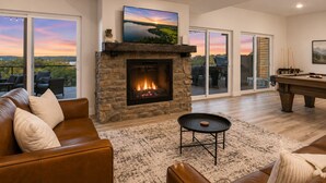 A living room with a stone fireplace, a pool table, and a large window overlooking a scenic landscape. - Hot Tub, Lake & Mtn Views, King Beds, Bunk Room (Kimberling City)