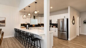 A modern kitchen with a central island, bar stools, stainless steel appliances, pendant lighting, and a view into the dining area. - Hot Tub, Lake & Mtn Views, King Beds, Bunk Room (Kimberling City)