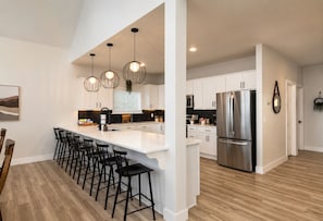 Una cocina moderna con una isla central, taburetes de bar, electrodomésticos de acero inoxidable, iluminación colgante y vistas al área de comedor.