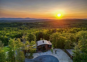 Ein modernes Haus mit gewölbtem Dach, umgeben von dichtem Wald, mit freiem Blick auf den Sonnenuntergang über sanften Hügeln.