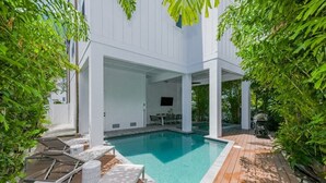 A modern outdoor pool area with a wooden deck, lounge chairs, and a dining area. - Mariner's Crossing – Luxury Escape w/ Rooftop Deck, Pool, Spa & Game Room (Bradenton Beach)