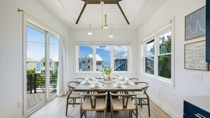 A dining area with a large table set for six, wooden chairs, and a view of the outside through large windows. - Mariner's Crossing – Luxury Escape w/ Rooftop Deck, Pool, Spa & Game Room (Bradenton Beach)
