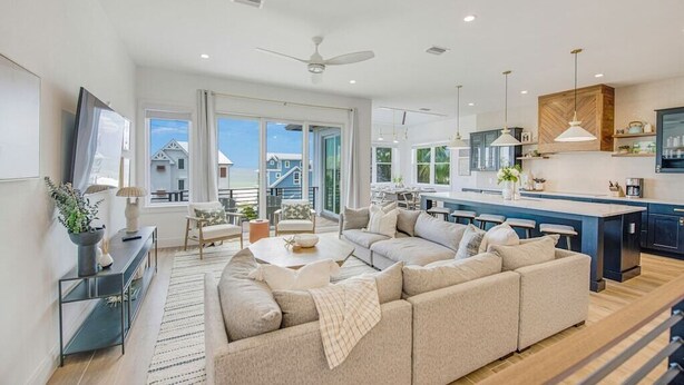 A modern living room with a large beige sectional sofa, a coffee table, and a TV on a stand. There is a dining area with a table and chairs, and a kitchen with a large island and pendant lights. - Mariner's Crossing – Luxury Escape w/ Rooftop Deck, Pool, Spa & Game Room (Bradenton Beach)