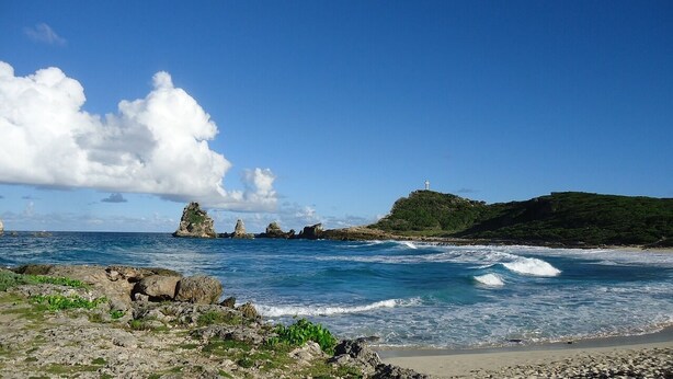 Un paisaje costero con terreno rocoso, una playa de arena y una cruz en una colina.