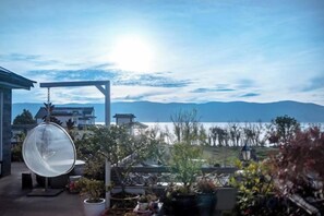 Une terrasse agrémentée d’un fauteuil suspendu, de plantes en pot et offrant une vue sur un plan d’eau et des montagnes.