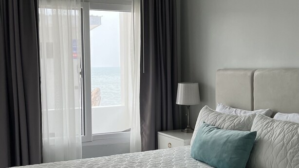 A hotel room with a large bed, a nightstand with a lamp, and a view of the sea through the window. - Classy Queen w/ramp access. Balcony subject to the sound of waves (La Ceiba)