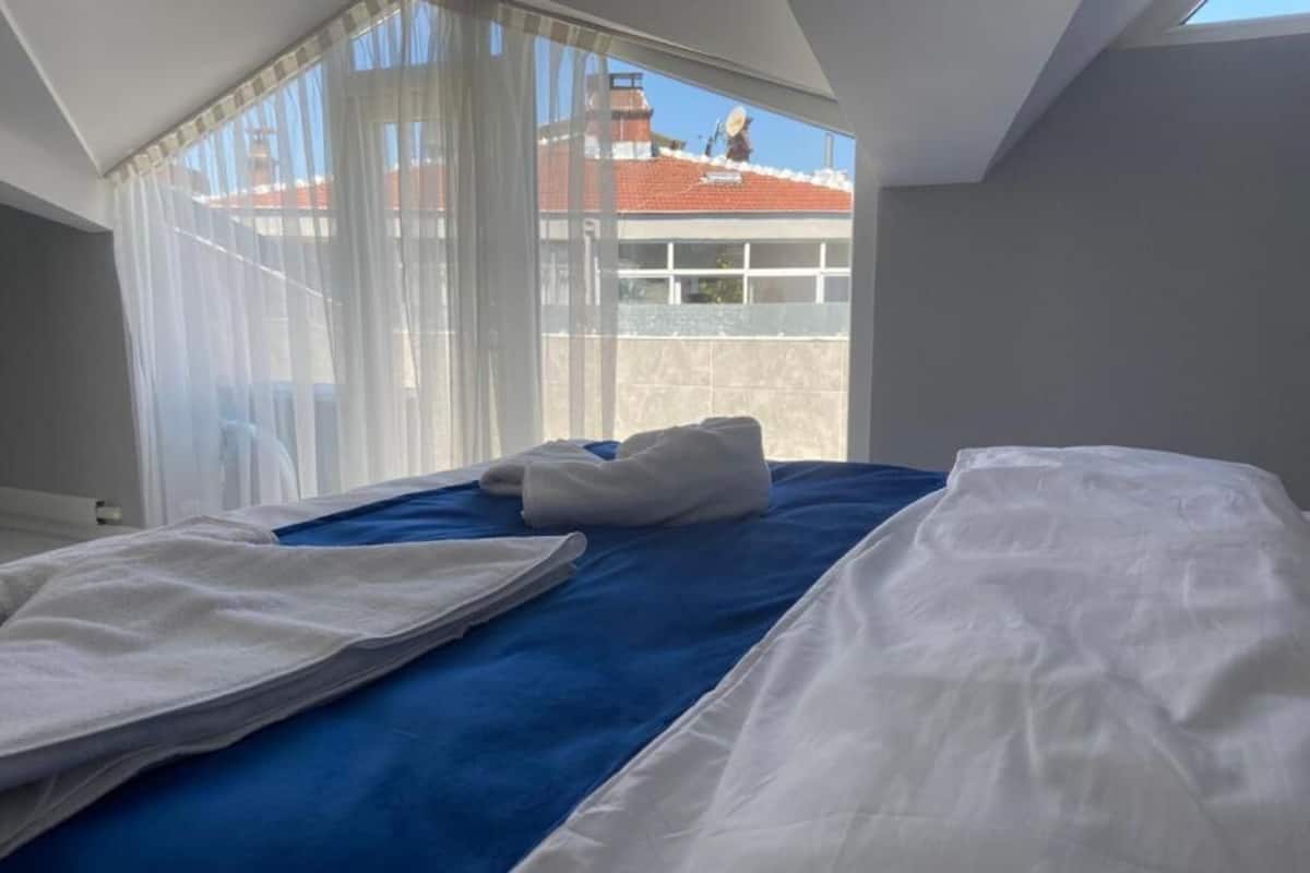 A bedroom with a bed, a window with sheer curtains, and a view of a building with a red roof.