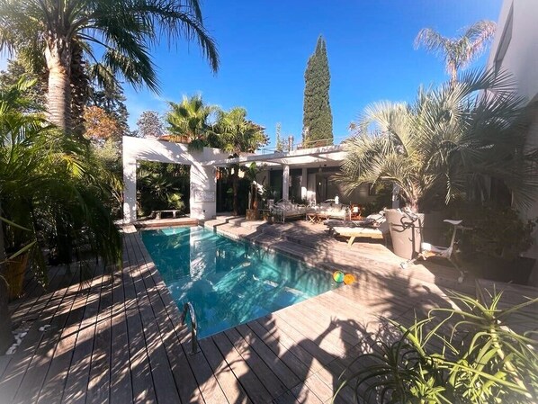 A swimming pool with a wooden deck, surrounded by palm trees and a tall evergreen.
