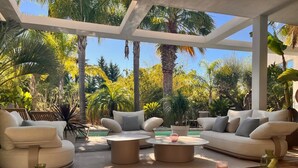Uma área de estar coberta ao ar livre com piscina, rodeada por plantas tropicais e palmeiras.