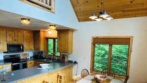 Una cocina moderna con armarios de madera, una isla central con taburetes, una mesa de comedor con sillas y un amplio ventanal con vistas a la vegetación.