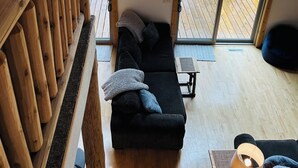 Interior acogedor de una cabaña con una terraza de madera, un sofá negro con cojines, una mesita y una lámpara.