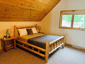 Ein hölzernes Bett mit Kopfteil, ein Nachttisch, eine Lampe und ein Fenster mit Blick auf Grünfläche.
