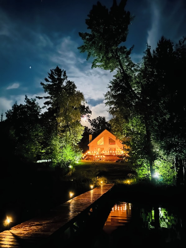 A house with a wooden deck and a pond in front, surrounded by trees.