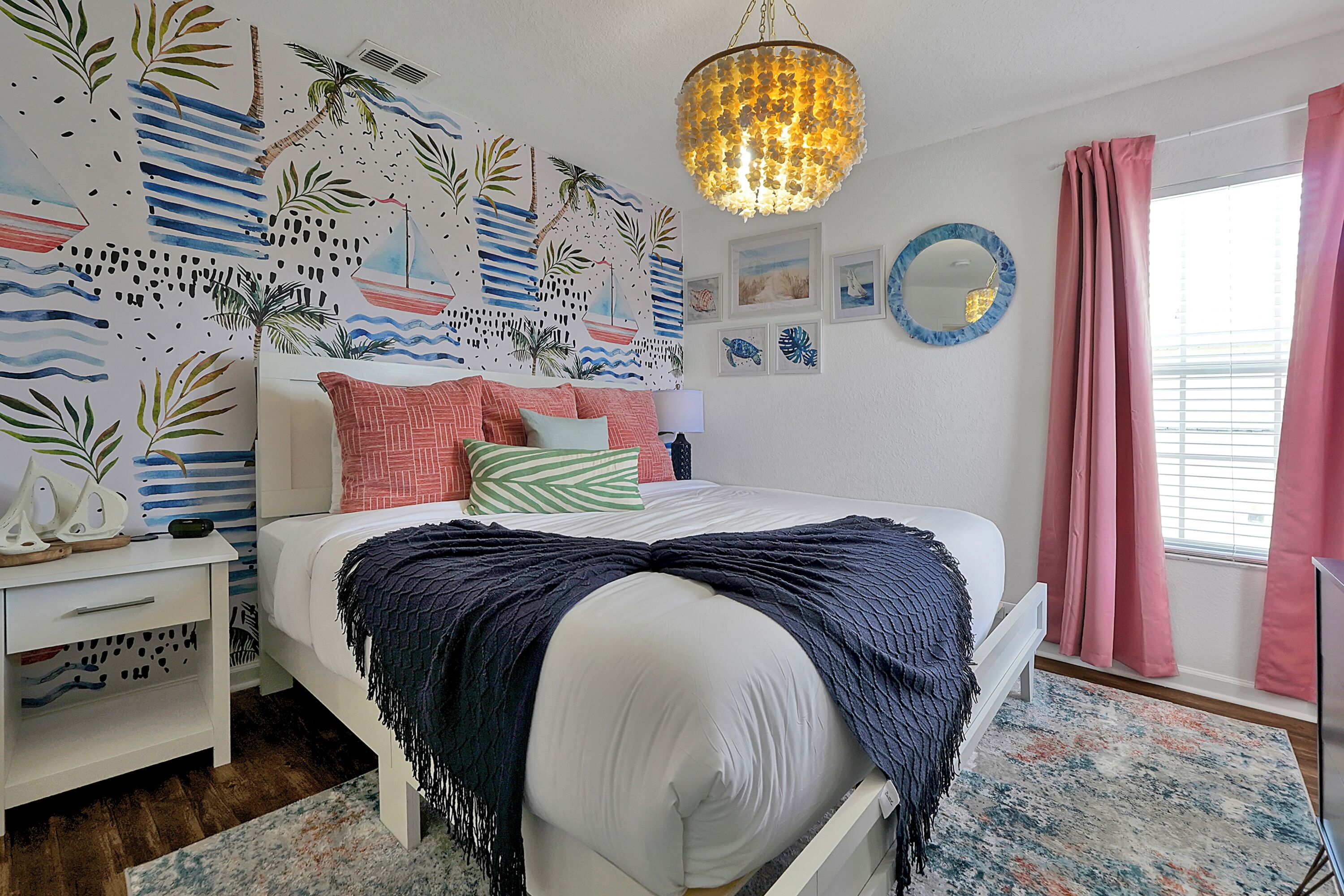 Un dormitorio con cama, mesita de noche, cómoda, candelabro, ventana con cortinas rosadas y un mural tropical en la pared.