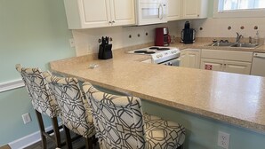 Una cocina con armarios blancos, estufa, microondas y cafetera. Hay tres taburetes de bar con diseños y una silla de madera.