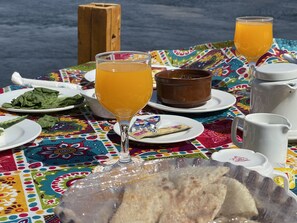 Ein Tisch ist gedeckt mit einer bunten Tischdecke, einem Glas Orangensaft, einem Teller mit grünen Blättern, einer Schüssel, einer Packung Snacks, einer weißen Tasse und einem Metallbehälter mit Fladenbrot.