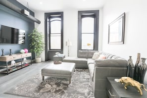 Een moderne woonkamer met een grijze bank, een bijpassend poufje, een flatscreen-televisie aan de muur, een kamerplant en een kamerbreed tapijt met een patroon.