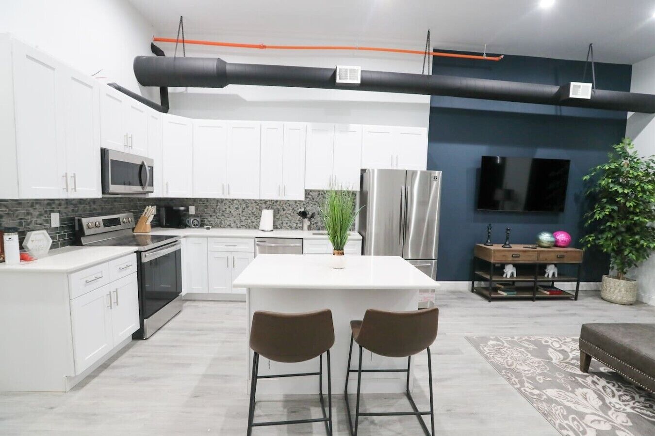 A modern kitchen with white cabinets, stainless steel appliances, a central island with two chairs, and a potted plant.