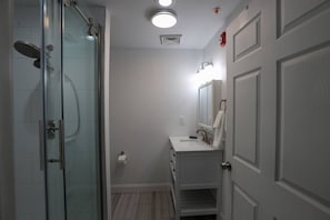 Een badkamer met een glazen douchecabine, een witte wastafel, een spiegel en een deur waarvan een handdoek aan hangt.