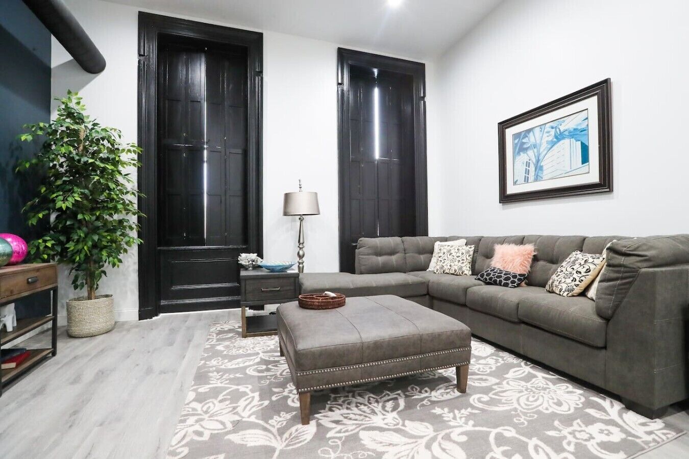 A living room with a grey sofa, a side table, a lamp, a potted plant, and a framed picture on the wall.