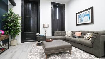 A living room with a grey sofa, a side table, a lamp, a potted plant, and a framed picture on the wall.