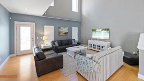 A spacious living room with a high ceiling, a large window, a flat-screen TV, and a sofa set.