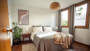 Un dormitorio con cama, mesitas de noche, lámpara, planta en maceta y ventana con cortinas.