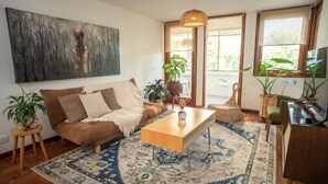 Sala de estar con un sofá marrón, una mesa de centro de madera y un tapete estampado. Hay plantas, una silla de mimbre y un televisor sobre un mueble.
