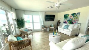 A spacious living room with a white sofa, a wicker chair, and a white armchair. There is a ceiling fan, a round coffee table, and a large window with curtains. - Panoramic view of the sugar white sand and the sparkling Emerald waters (Port St. Joe)