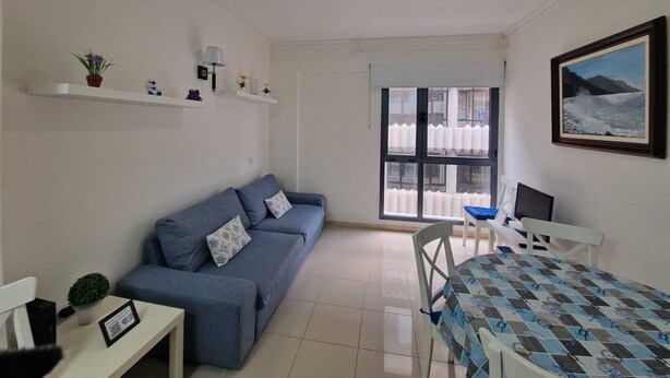 Una habitación con sofá, una mesa con mantel estampado, una silla, un escritorio con computadora, estantes con adornos y un cuadro en la pared.